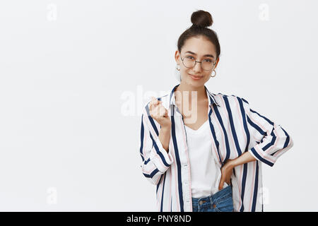 Ich habe alles unter Kontrolle. Studio geschossen von zuversichtlich charmante weibliche in Gläsern und Shirt mit bun Frisur und deutete mit der Hand und hält Palm auf Taille, grinsend, selbstbewusst und glücklich Stockfoto