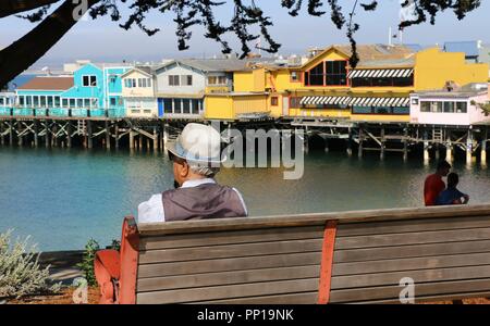 Ein älterer Mann mit einem Adretten Hut sitzt auf einer Bank mit Blick auf Fisherman's Wharf, Monterey, CA. Stockfoto