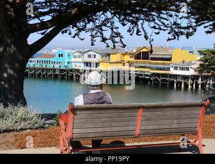 Ein älterer Mann mit einem Adretten Hut sitzt auf einer Bank mit Blick auf Fisherman's Wharf, Monterey, CA. Stockfoto