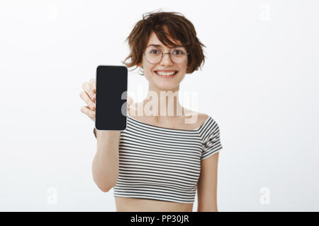 Sehen Sie, es ist mein Profil in neuen sozialen Netzwerk. Portrait von positiven gut aussehende urban Frau in runde Gläser, Ziehen von Hand mit Smartphone in Richtung Kamera, zeigt neue App oder Foto über graue Wand Stockfoto