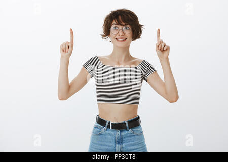 Mädchen mag auf die Sterne zu schauen. Verträumt attraktive Frau mit kurzen braunen Haaren in runde Gläser und top Gestreifte 7/8, nach oben gerichtet mit erhobenem Zeigefinger und breit grinsend, während neben den Blick Stockfoto