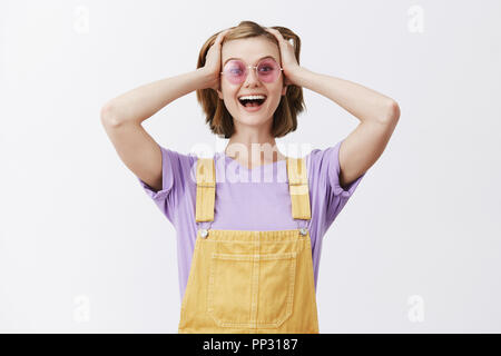 Nein, Du kidding mich. Überrascht von Freude und gut aussehenden Studentin in der Trendfarbe gelb Latzhose und Sonnenbrille aufgeregt, die Hände auf den Kopf und breit grinsend und sorglos an Kamera Stockfoto