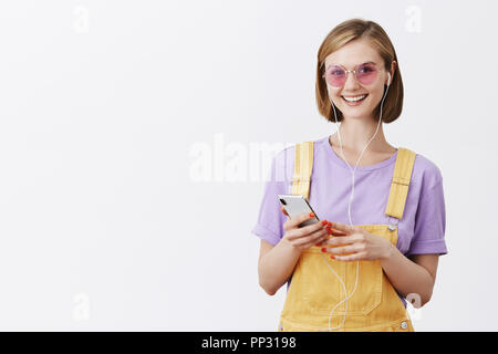 Mädchen fertig, von zu Hause aus zu Fuß gehen, Ohrhörer und Kommissionierung positiver Song, stehend gelb Latzhose und Sonnenbrille, Smartphone und breit grinsend an Kamera Stockfoto