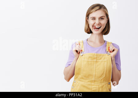 Zeit, etwas Spaß zu haben. Charmante sorglos und sicher flirty Freundin Latzhose ziehen und freudig winking und zeigt Zunge und hinting Freund über anstehende Party, immer bereit zu Rock Stockfoto