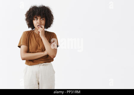 Innen- schuss auf zweifelhafte und befragt thoghtful junge afrikanische amerikanische Frau mit Afro Frisur in Braun t-shirt beißen Fingernagel und stirnrunzelnd blickt nach rechts, während die Entscheidung oder Denken Stockfoto