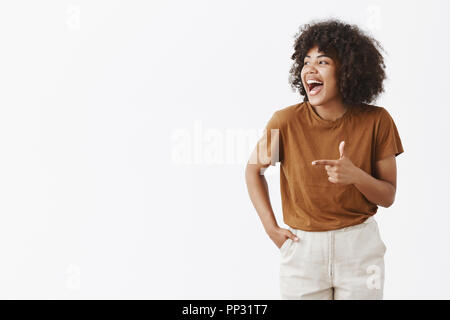 Nicht das wahre Glück verbergen. Portrait von fröhlichen charmanten Sorglos afrikanische amerikanische Frau mit Curly Frisur suchen und zeigt mit dem Finger Gewehr Lachen vom Glück und gute Laune links Stockfoto