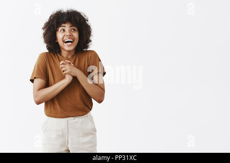 Studio shot von aufgeregt und verträumt schön fit African American female mit Afro Frisur lacht freudig Holding Palmen in der Nähe von Herz und und schaut von Glück und Freude über graue Wand Stockfoto