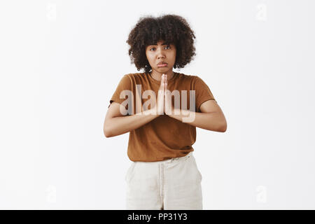 Studio shot düsterer umgekippt gut aussehende dunkelhäutigen Teenager Frau mit Afro Frisur schürzte die Lippen in trauriges Lächeln runzelte die Stirn und Hände halten im Gebet fragen zu helfen oder über graue Wand gefallen Stockfoto