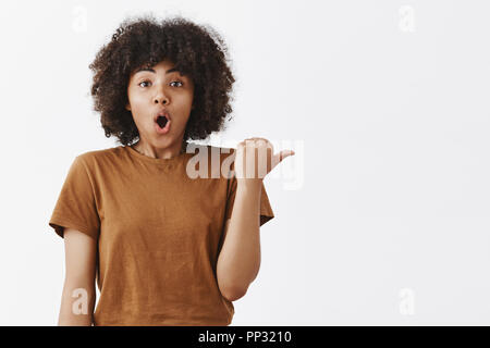 Aufgeregt und betäubt beeindruckt dunkelhäutige Mädchen miteinem afro Frisur in braun Trendy T-shrit Falten öffnete die Lippen nach rechts mit Daumen Beschreibung super Szene sah sie, daß Richtung Stockfoto