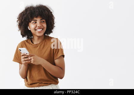 Studio shot sorglosen freundliches und kreatives stilvolle afrikanische amerikanische Teenager in Braun t-shirt rechts drehen mit breiten zufriedenen Lächeln tragen drahtlose Kopfhörer und Holding smartphone Stockfoto