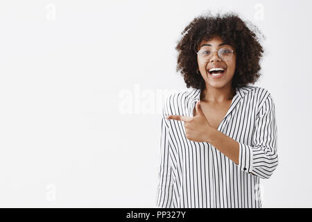 Über-emotional aufgeregt und glücklich niedlichen afrikanische amerikanische Frauen in gestreifte Bluse und Gläser Lachen aus Spaß und Freude mit Zeigefinger in grosse Zeit über graue Wand amüsiert Links Stockfoto