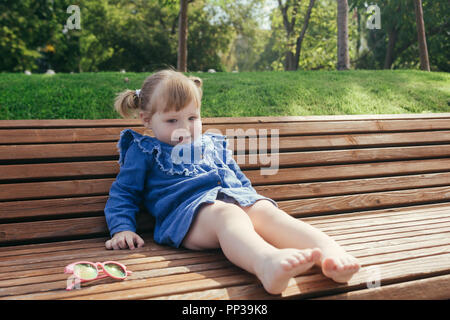 Kleines Mädchen auf der Bank sitzen in einem Stadtpark an einem warmen sonnigen Tag Stockfoto