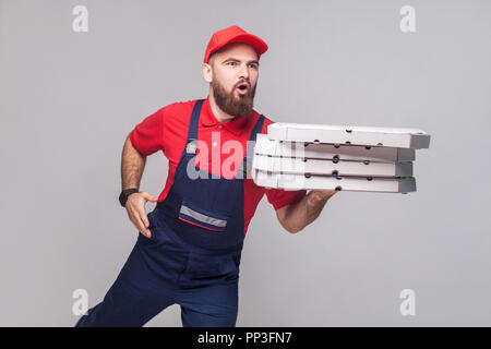 Beeilen Sie sich! Junge erstaunt Lieferung Mann mit Bart in der blauen Uniform und dem Roten t-shirt Holding stack Pizzakartons und ausführen, um die Bereitstellung auf grauen Hintergrund. Ich Stockfoto