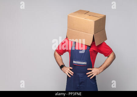 Porträt der jungen Logistik Lieferung Mann in der blauen Uniform und dem roten T-Shirt stehen und das Gesicht mit offenen Karton auf grauem Hintergrund, Hände o Stockfoto