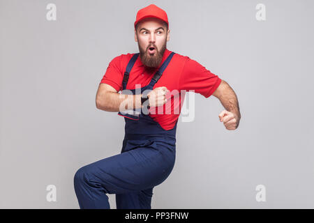 Beeilen Sie sich! Junge erstaunt Heimwerker mit Bart in blauen, roten T-Shirt und Kappe sind spät dran und Starten für Hilfe auf Zeit zu laufen. Grauer Hintergrund, Hallenbad Stockfoto