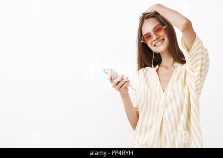 Richtige Lied in den Ohren macht Tag heller. Sorglos wunderschöne junge stilvolle Frau, Sonnenbrille, gelbe Bluse berühren Frisur Neigekopf freudig Holding Smartphone Musik im Kopfhörer. Stockfoto