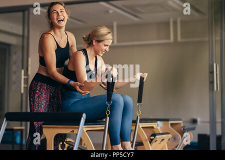 Lachend weiblichen Trainer helfen, eine Frau in Ziehen stretch Bands sitzen auf Pilates Training Maschine. Lächelnde Frau an der Turnhalle tun Pilates Training Stockfoto