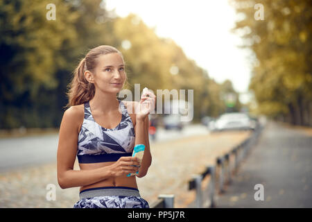 Sportlich attraktive schlanke junge Frau sitzt auf einem strassenrand Rail essen ein Stück von einem Protein Bar, als sie eine Pause von ihren täglichen Jog nimmt Stockfoto