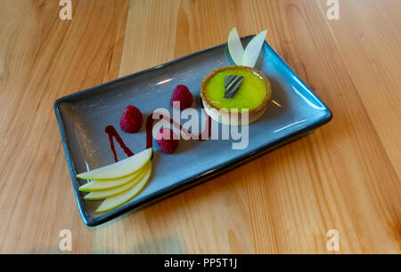 Ein Key Lime tart mit ein paar Himbeeren und in Scheiben geschnittenen Apfel auf eine rechteckige, blaue Schale Stockfoto