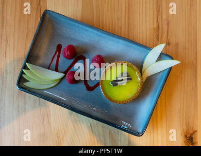 Ein Key Lime tart mit ein paar Himbeeren und in Scheiben geschnittenen Apfel auf eine rechteckige, blaue Schale Stockfoto