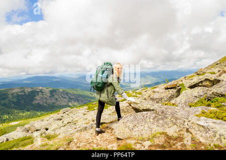 Junge Mädchen reisen mit großen grünen Rucksack und Flasche, entlang der grünen Hügel mit grünem Gras setzt auf einen großen Stein für Entspannung unter den blauen Himmel Stockfoto