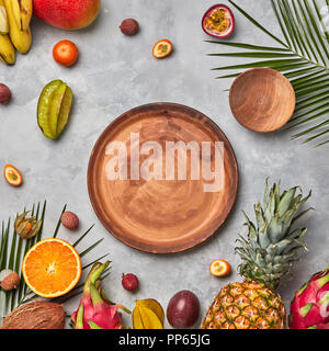 Verschiedene saftige exotische Früchte, Kokos, Litschis, Carom, Ananas, Palmenblättern und leeren braunen Holz- Platten auf grauem Beton Hintergrund mit Platz für Text. Flach Stockfoto