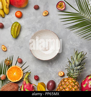 Ganze oder halbe exotische gesunde Früchte, Karambolen, Ananas, Passionsfrucht, Pitahaya, Palm grüne Blätter und einen leeren Teller auf einem grauen Beton Hintergrund mit Platz für Text. Flach Stockfoto