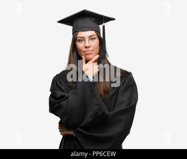 Junge Hispanic Frau tragen graduierte Cap und einheitliche schaut zuversichtlich in die Kamera mit einem Lächeln mit verschränkten Armen und Hand am Kinn angehoben. Denken Stockfoto