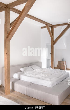 Modernes Schlafzimmer mit weißen Wänden, Holzbalken und Parkettboden mit Teppichen auf dem Boden. Es ist ein grauer Bett mit Licht Handtücher, Truhen mit einer Statuette, Stockfoto