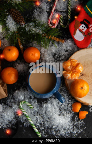 Weihnachten und Neujahr Konzept. Mandarinen, Kaffeebecher, Schnee, Weihnachten Baumstrukturzweig. Vor einem dunklen Hintergrund. Flach Top View Stockfoto