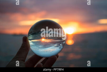 Helles Orange Marine Sonnenuntergang mit Welle und Sonne am Horizont gefangen in der Reflektion im Glas Kugel gehalten in den Fingerspitzen Stockfoto