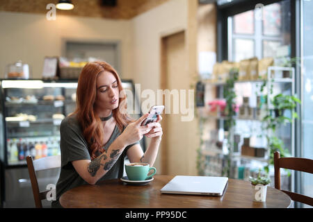 Red Head Mädchen mit Arm Tattoo, allein in einem Cafe an Ihr Mobiltelefon suchen sitzend, mit einem Laptop saß auf dem Holztisch in Liverpool, Großbritannien Stockfoto