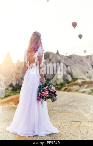 Frau in einem langen Kleid auf dem Hintergrund von Luftballons in Kappadokien. Mädchen mit Blumen Hände steht auf einem Hügel und blickt auf eine große Zahl von fliegenden Ballons Stockfoto