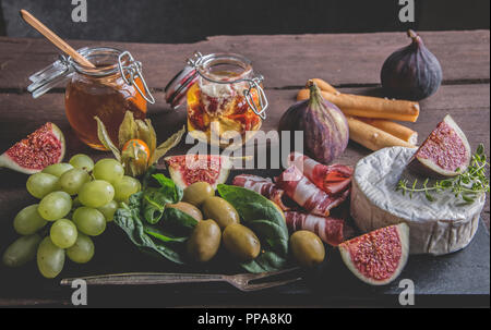 Prosciutto, Camembert, Feigen, Spinatblätter, Honig, Trauben, Oliven auf dunklen, das Brett über rustikal Hintergrund Stockfoto