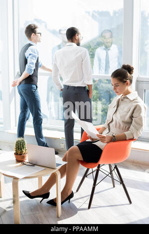 Volle Länge Portrait von eleganten Geschäftsfrau Lesen von Dokumenten während der Arbeit in modernen Büro durch Sonnenlicht mit zwei Personen im Hintergrund leuchtet Stockfoto