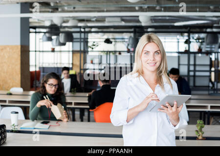 Taille bis Portrait von Blond moderne Geschäftsfrau lächelnd an Kamera und halten Digitale tablet Während in Open Space Office posing Stockfoto