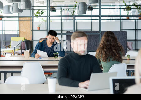 Gruppe von Personen, die in der Open Space Office, Schwerpunkt auf jungen asiatischen Mann Lesen von Dokumenten in zurück Stockfoto