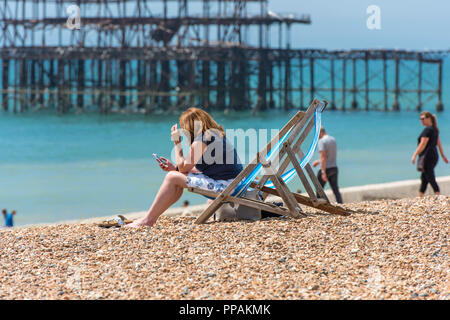 Frau ihr Mobiltelefon während der Sitzung in einem gestreiften Liegestuhl am Strand in Brighton, gegenüber der niedergebrannt Pier. Stockfoto