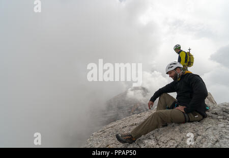 Junge männliche Bergsteiger auf einem Berggipfel der Dolomiten genießen die Aussicht vom Gipfel und seine Bergführer stehen hinter ihm Stockfoto