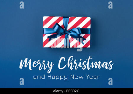 Geschenkbox rot gestreifte Papier gewickelt und mit blauer Schleife auf dunkelblauem Hintergrund gebunden. Weihnachtskarte mit Aufschrift frohe Weihnachten. Stockfoto