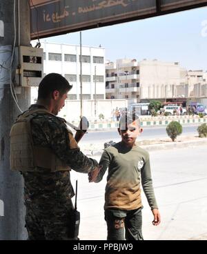 Eine Manbij militärischen Rat Mitglied schüttelt Hände mit einem lokalen Kind während des Eid al-Adha, was übersetzt "Festival der Opfer "in Manbij, Syrien, 21. August 2018. Wenn die Sicherheit in der Stadt, MMC-Mitglieder haben die Möglichkeit einer Beziehung mit der lokalen Bevölkerung zu errichten und die Unbedenklichkeit und Sicherheit der Bereich gewährleisten. Stockfoto