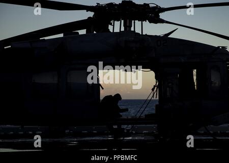 Ein Seemann mit Helikopter Meer Combat Squadron 25 sitzt in einem U.S. Navy MH-60S Seahawk Hubschrauber auf dem Flugdeck der Amphibisches Schiff USS Wasp (LL 1), wie die Sonne vor der Küste von Okinawa, Japan, August 25, 2018. HSC-25 führt kritische Unterstützung für Marine Corps und Marine amphibische Operationen während der Fahrt als Teil der Wasp Amphibious Ready Gruppe. Marinesoldaten und Matrosen mit der 31 Marine Expeditionary Unit, bestehend aus einem Befehl Element, einem Bodenkampf Element, eine Aviation Combat Element und eine Logistik Combat Element, an Bord der Schiffe der US-Marine die Wasp AR Stockfoto