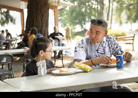 U.S. Army Staff Sgt. Isaac Kumia, eine menschliche Ressourcen noncommissioned Officer mit der 104-Ausbildungszentrum (LT), Sitz und Hauptverwaltung, Aktien sein Mittagessen mit seinem Kind während der organisierten Familie seiner Einheit - Tag 12.08.19, 2018 in Lakeside Park auf Joint Base Lewis-McChord, Washington der US-Armee finden Einheit aufgefordert, seine Truppen und ihrer Familien, die gemeinsam für die Lakeside Ereignis, gegrillten Speisen, Spiele im Freien und militärische Ressourcen Aufbau an Tabellen enthalten. Stockfoto