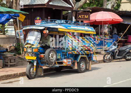 Treiber in einem geparkten bunten Dreirad Taxi genannt Jumbo (oder Tuk Tuk) auf dem Phetsarat Straße in Luang Prabang, Laos, an einem sonnigen Tag. Stockfoto