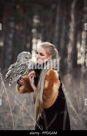 Frau blond im Herbst im Pelzmantel mit Eule an Hand der erste Schnee. Schöne Mädchen mit langen Haaren in der Natur, eine Eule. Romantisch, zarten look Mädchen Stockfoto