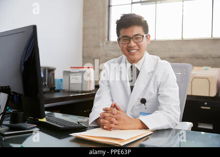 Jungen Asiatischen männlicher Arzt am Schreibtisch sitzen, Porträt Stockfoto