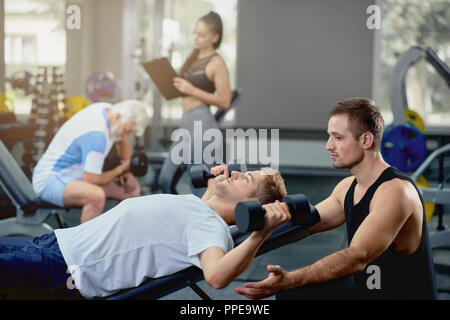 Junge freundliche Personal Coach helfen, muskulöser Kerl, Übungen mit Hanteln in dunklen Turnhalle durchführen. Auf unscharfen Hintergrund, heißes Mädchen, Personal Trainer, Unterstützung von älteren grauen Mann. Stockfoto