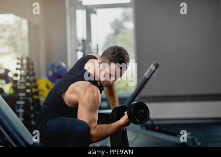 Schöne sportliche Mann verbiegen Muskeln mit Barbell in dunklen Fitnessraum. Sport Konzept. Atheletic muskulöse Kerl tragen Sport Hosen und schwarze Jersey, trainieren, während in dunklen Turnhalle sitzen. Konzept der Sport. Stockfoto
