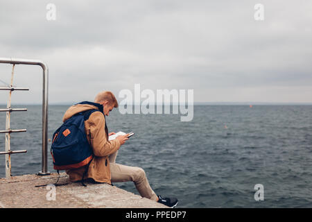 Student mit Rucksack lesen Buch von Meer auf Pier in Odessa an regnerischen Herbsttag. Guy studieren im Freien Stockfoto