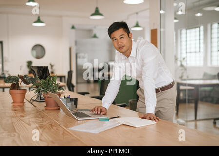 Junge asiatische Geschäftsmann lehnte sich auf seinem Schreibtisch mit einem Laptop Stockfoto